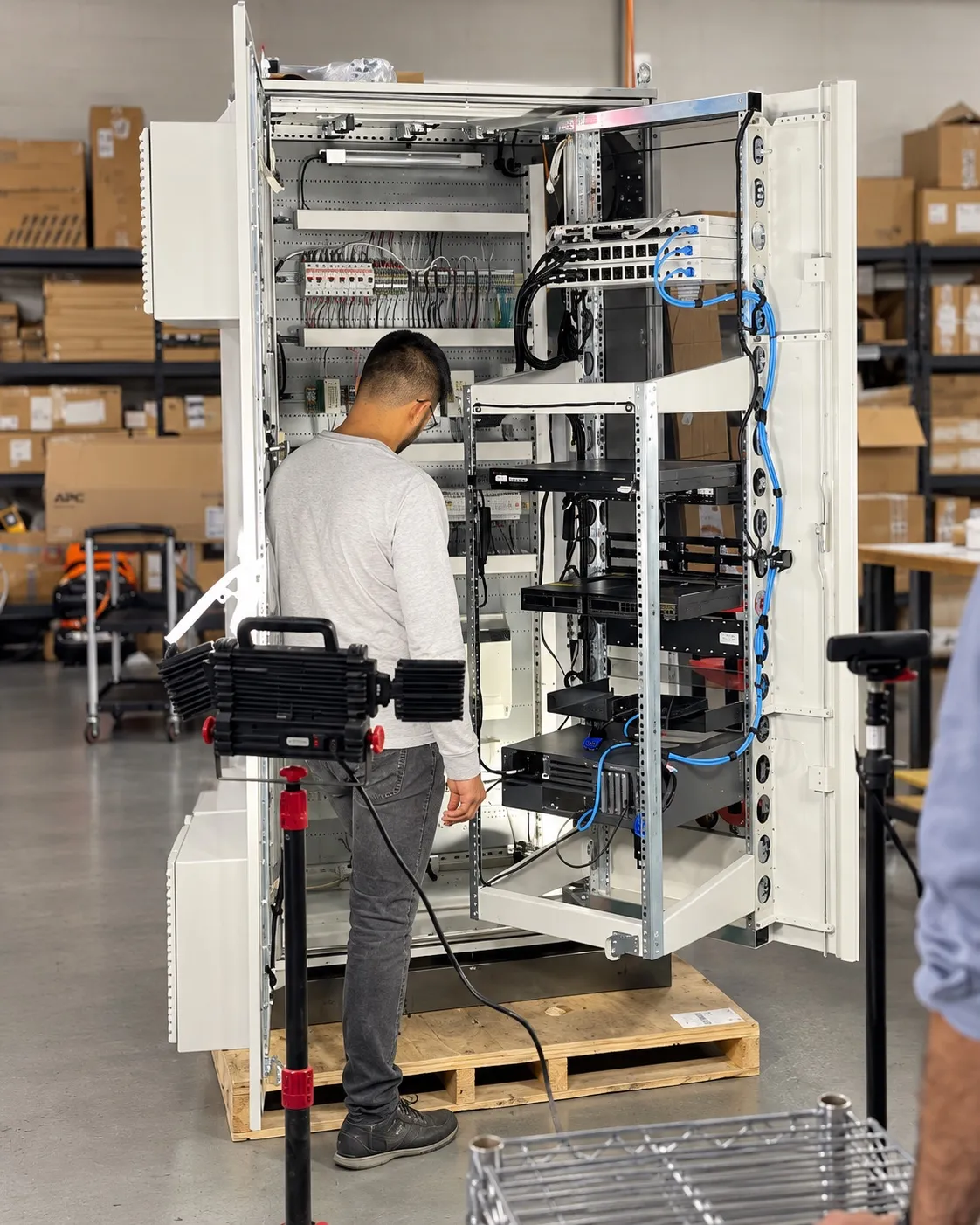 Technician working on an open communication cabinet with rack-mounted network switches and blue patch cables in a workshop